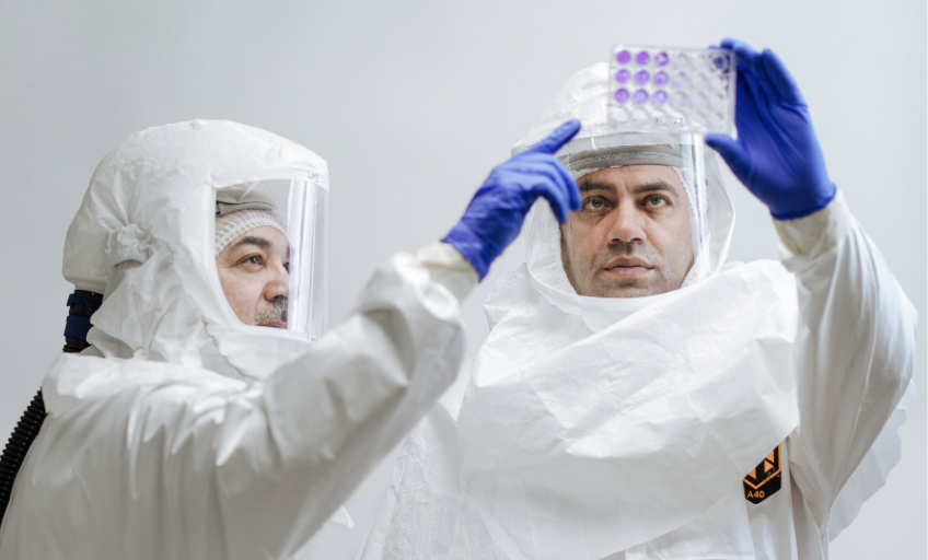 Two scientists in white protective suits, hoods and gloves looking at cell culture plate.